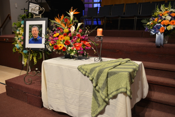 Display of framed photo of Ben Lam beside a table with a white tablecloth. On the table is a large arrangement of flowers, a lit candle, and a hand-knit blanket.