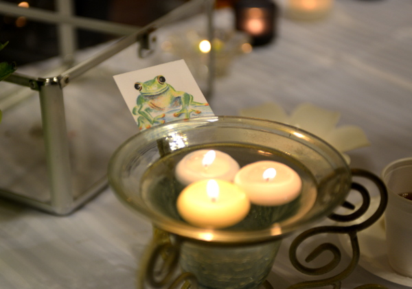 A glass stand with three floating candles that are lit, in front of a square magnet with a frog drawing on it.