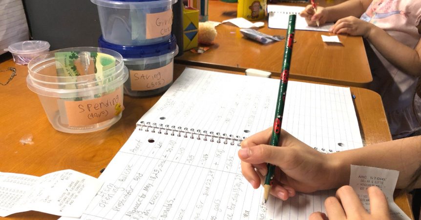 Close up of child holding a pencil writing in her allowance ledger. Also on the desk are three containers marked "Saving," "Spending," and "Giving".