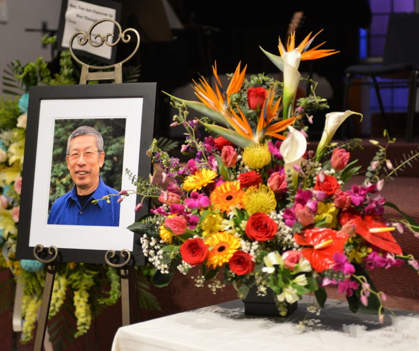 A framed photo of Ben Lam is displayed on an easel next to a large multicoloured floral arrangement displayed on a white table.